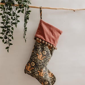 May include: A Christmas stocking with a floral pattern in shades of black, orange, and green. The top is a solid dusty rose color, trimmed with small brown pom-poms. It hangs from a wooden branch.