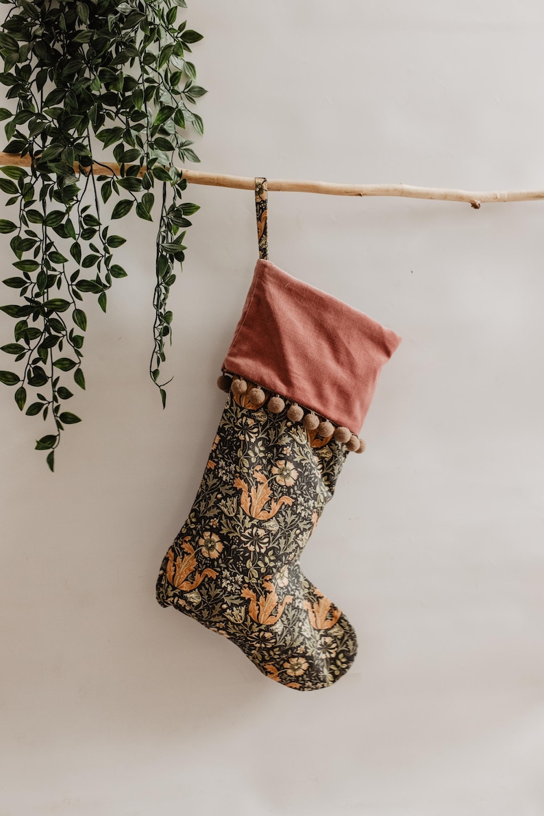 May include: A Christmas stocking with a floral pattern in shades of black, orange, and green. The top is a solid dusty rose color, trimmed with small brown pom-poms. It hangs from a wooden branch.