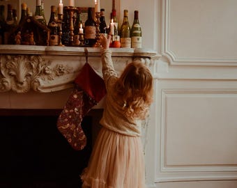 May include: A young person in a gold-colored sweater and peach-colored skirt reaches for items on a fireplace mantel. The mantel is decorated with bottles, candles, and a Christmas stocking. A chandelier hangs above.