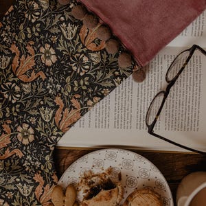 May include: A dark floral patterned fabric with a burgundy velvet edge and brown pom-poms. A plate of pastries and gingerbread cookies sits near an open book and eyeglasses.