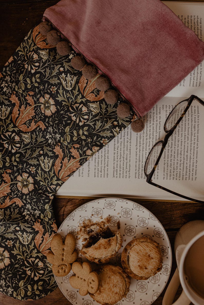 May include: A dark floral patterned fabric with a burgundy velvet edge and brown pom-poms. A plate of pastries and gingerbread cookies sits near an open book and eyeglasses.