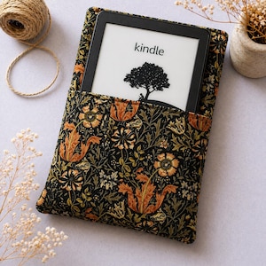 May include: A Kindle e-reader in a black case with a floral pattern. The case has a black base with orange, gold, and green floral designs. The Kindle screen shows the word "kindle" and a tree silhouette.