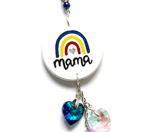 May include: A white circular charm with a rainbow design and the word "mama" in black script. Below, three heart-shaped crystals in blue, iridescent, and magenta dangle from chains. The charm is suspended by a beaded strand.