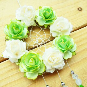 May include: A handmade dreamcatcher featuring a circular woven web, adorned with white and green fabric roses. The dreamcatcher has hanging beads and a green tassel, set against a wooden background.
