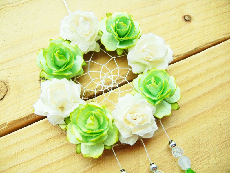 May include: A handmade dreamcatcher featuring a circular woven web, adorned with white and green fabric roses. The dreamcatcher has hanging beads and a green tassel, set against a wooden background.