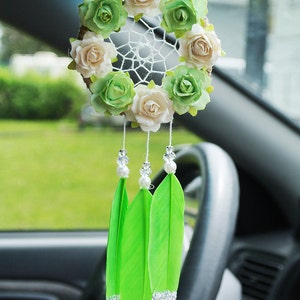 May include: A floral dreamcatcher with a woven web, adorned with cream and green fabric roses. Three bright green feathers with silver glitter tips hang below. A decorative car accessory.