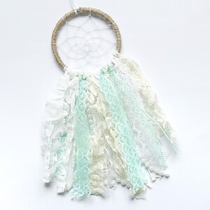 May include: A handmade dreamcatcher with a woven, twine-wrapped hoop and delicate white web. The dreamcatcher features flowing ribbons of white, cream, and pale blue lace, creating a soft, airy aesthetic.