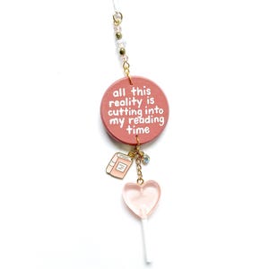 May include: A pink charm with the text "all this reality is cutting into my reading time." It features a book charm, a heart-shaped lollipop, and gold-colored beads. The charm hangs from a thin, clear string.