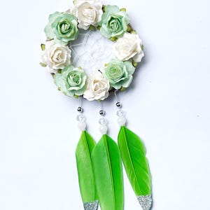 May include: A dreamcatcher with a circular woven frame adorned with white and green paper roses. Three bright green feathers with silver glitter tips hang below, accented with clear and white beads. A white string is at the top for hanging.