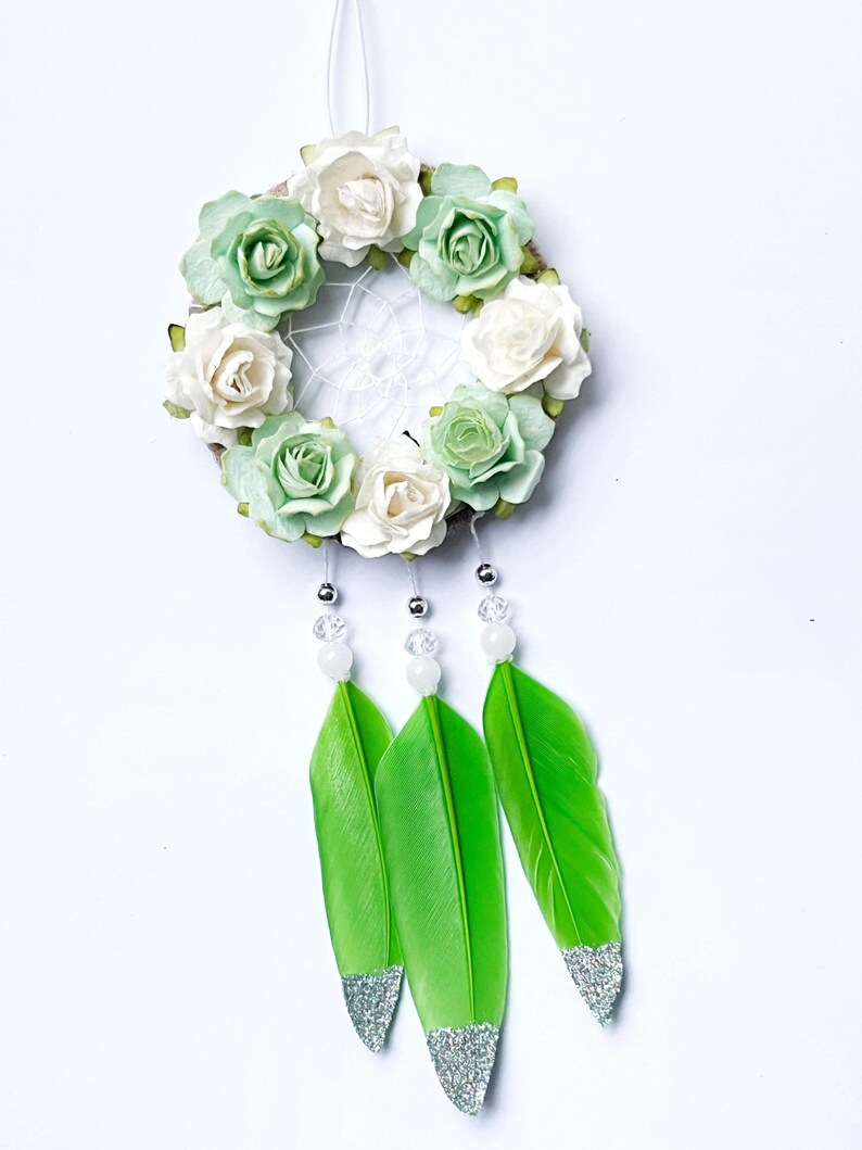 May include: A dreamcatcher with a circular woven frame adorned with white and green paper roses. Three bright green feathers with silver glitter tips hang below, accented with clear and white beads. A white string is at the top for hanging.