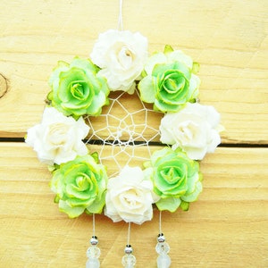 May include: A handmade dreamcatcher featuring a circular woven web, adorned with alternating white and green paper flowers. The flowers are arranged around the web's perimeter. Clear beads and silver accents dangle from the bottom, suspended against a wooden background.