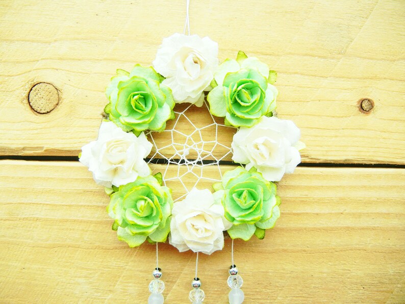 May include: A handmade dreamcatcher featuring a circular woven web, adorned with alternating white and green paper flowers. The flowers are arranged around the web's perimeter. Clear beads and silver accents dangle from the bottom, suspended against a wooden background.