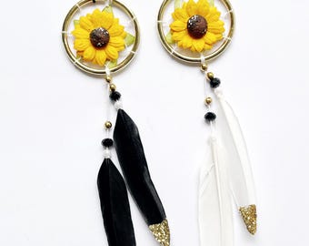 May include: Two dream catchers with gold-colored hoops, featuring yellow sunflower accents and black or white feathers with gold glitter tips. Each has a string for hanging, and decorative beads.