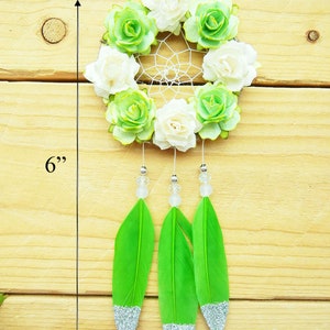 May include: A handmade dreamcatcher, approximately 6 inches long, featuring a circular woven web adorned with white and green fabric flowers. Three bright green feathers with silver glitter tips dangle below, accented with clear beads.
