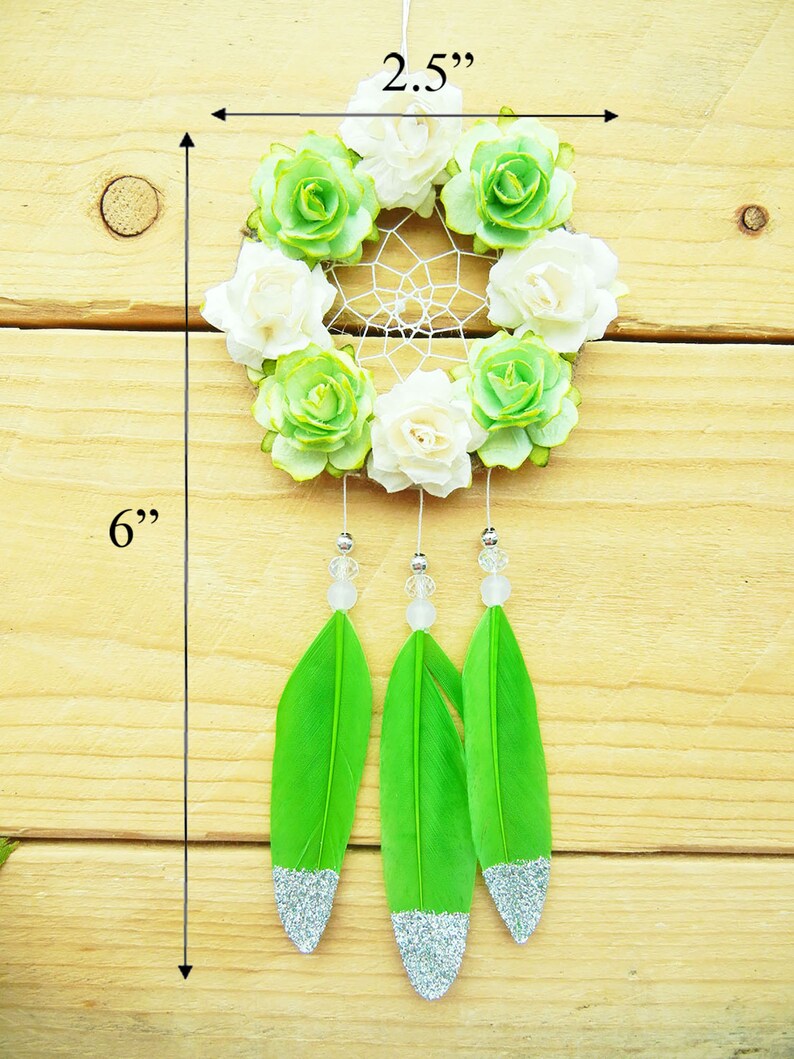 May include: A handmade dreamcatcher, approximately 6 inches long, featuring a circular woven web adorned with white and green fabric flowers. Three bright green feathers with silver glitter tips dangle below, accented with clear beads.