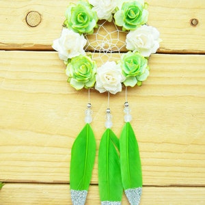 May include: A handmade dreamcatcher with a white web, adorned with white and green fabric flowers. Three bright green feathers with silver glitter tips hang below, accented with clear beads. The dreamcatcher is suspended against a light wood background.