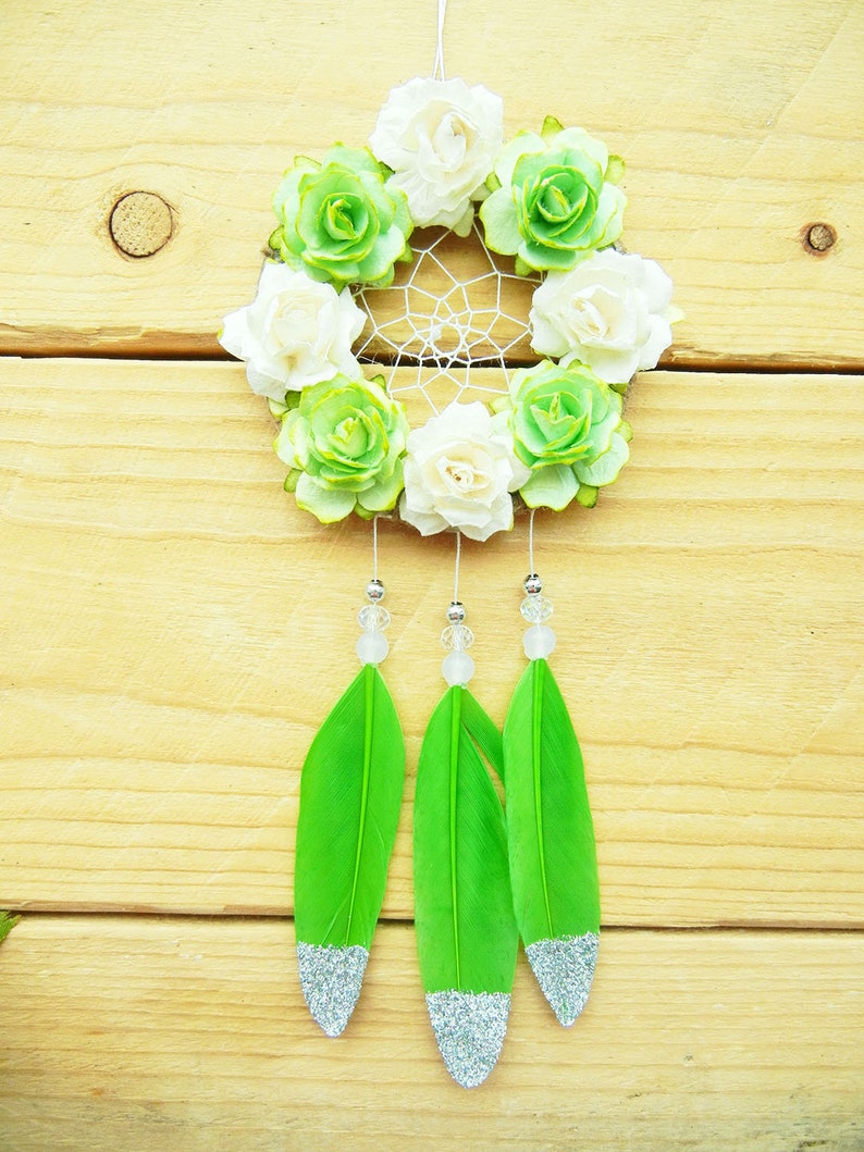 May include: A handmade dreamcatcher with a white web, adorned with white and green fabric flowers. Three bright green feathers with silver glitter tips hang below, accented with clear beads. The dreamcatcher is suspended against a light wood background.