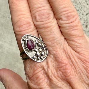 May include: A silver ring with a large, pear-shaped, deep red gemstone at the center. The ring has a textured, oval-shaped face with smaller, round silver accents. The band features a double-ring design.