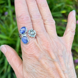 May include: A silver ring with two oval blue gemstones and a flower design. The ring is worn on a person's finger.