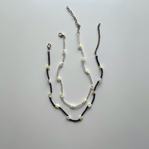 May include: Two delicate necklaces with daisy flower charms. One necklace is black and the other is silver.