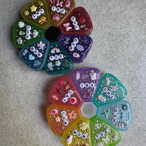 May include: Two circular pill organizers shaped like flowers, each with seven wedge-shaped compartments labeled with the days of the week. The lids are decorated with various small colorful charms, beads, and white lettered beads for each day.