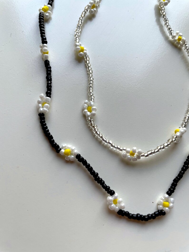 May include: Two necklaces with white daisy flower beads. One necklace has black beads and the other has clear beads.