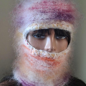May include: A colorful, hand-knitted balaclava in shades of pink, purple, orange, and white. The face opening is framed with a white and gold trim. The balaclava is made of a soft, fuzzy yarn, and is displayed on a mannequin head.