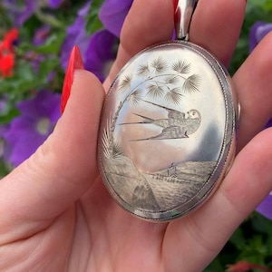 May include: Oval silver locket featuring an engraved bird in flight over a landscape with a tree. The locket has a detailed border and a loop for a chain. The background features purple flowers.