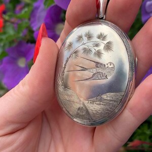 May include: Oval silver pendant with an engraved landscape scene featuring a bird in flight, a tree, and a small figure. The pendant has a detailed, textured border and a loop for a chain.