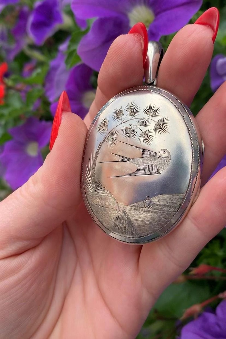 May include: Oval silver pendant with an engraved landscape scene featuring a bird in flight, a tree, and a small figure. The pendant has a detailed, textured border and a loop for a chain.