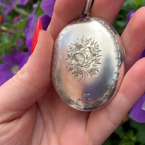 May include: Oval silver locket with a floral design. The locket has a scalloped edge and a loop for a chain. The front of the locket features an engraved rose and leaf pattern. The locket is held in a hand with red nail polish.
