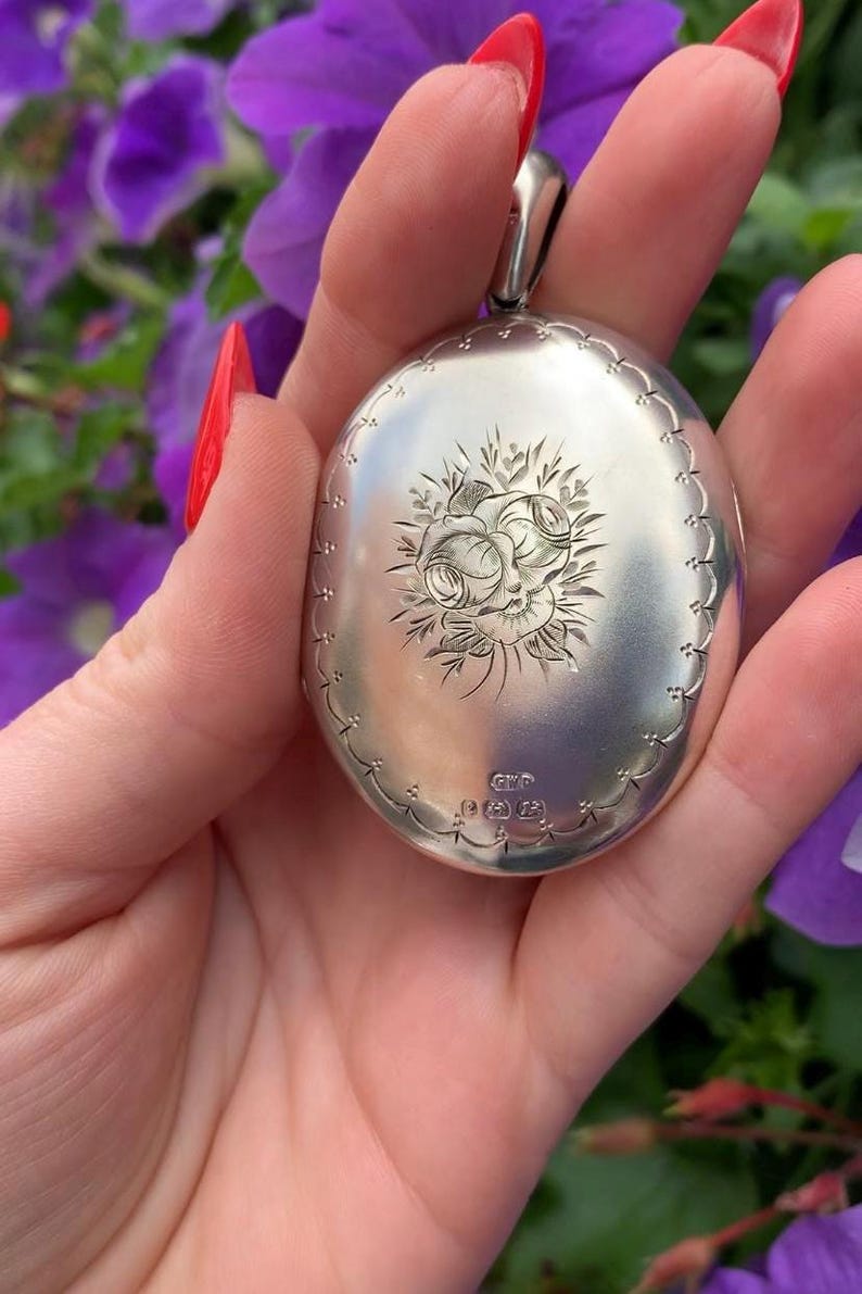May include: Oval silver locket with a floral design. The locket has a scalloped edge and a loop for a chain. The front of the locket features an engraved rose and leaf pattern. The locket is held in a hand with red nail polish.