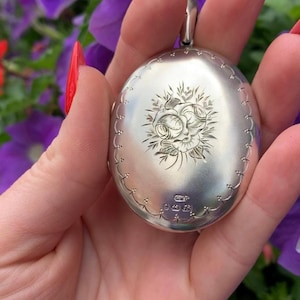 May include: Oval silver locket with a floral engraving. The locket has a decorative border and a loop for a chain. The background features purple flowers. The locket is likely made of sterling silver.