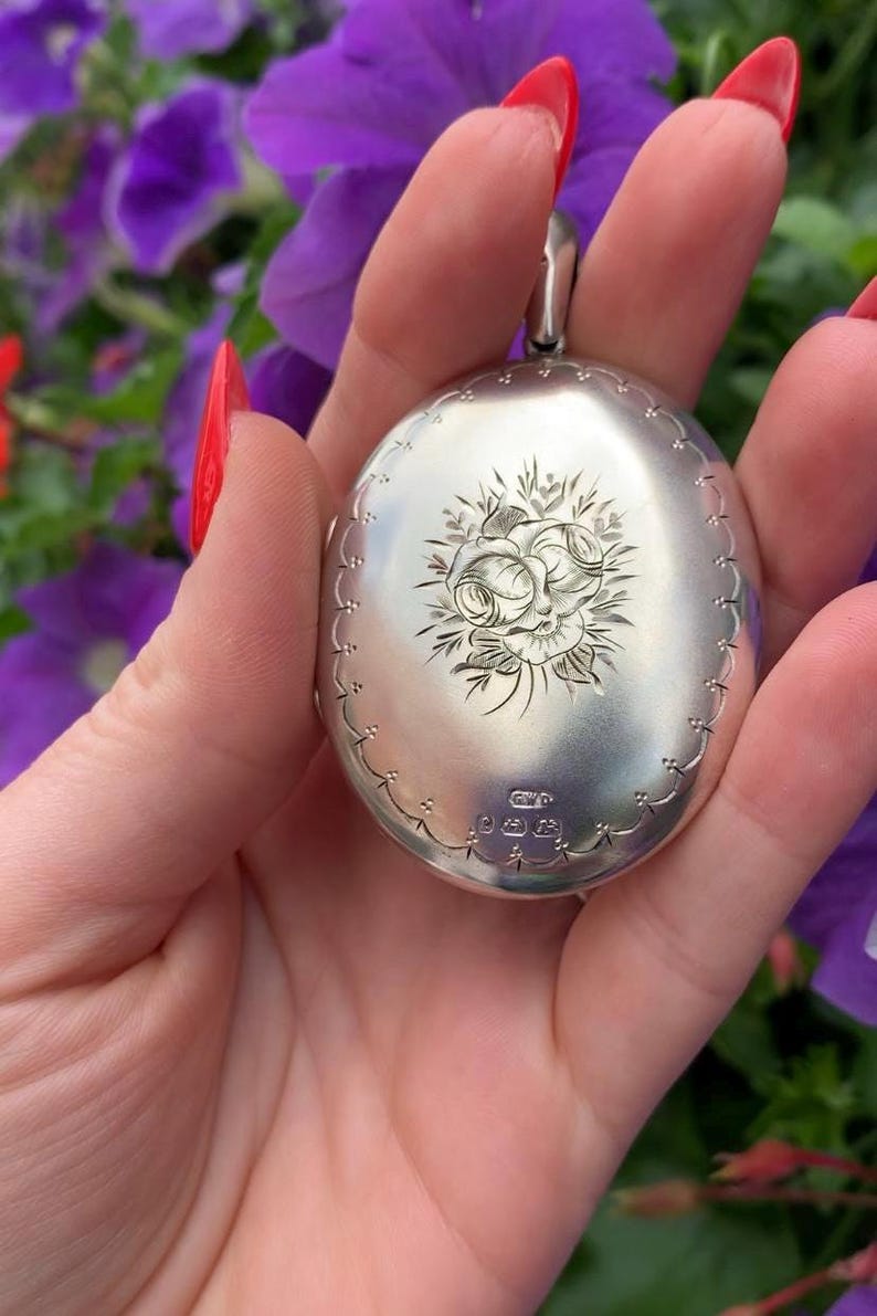 May include: Oval silver locket with a floral engraving. The locket has a decorative border and a loop for a chain. The background features purple flowers. The locket is likely made of sterling silver.