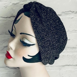 Pode incluir: Um turbante preto com um brilho metálico sutil, exibido em uma cabeça de manequim com traços pintados. O turbante é feito de um tecido macio e texturizado e tem um detalhe de nó franzido na parte superior. O fundo é uma parede de tijolos brancos.