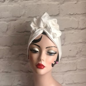 Może przedstawiać: Biały turban w stylu turbanu z dużą kokardą na górze. Owijka wykonana jest z miękkiej, elastycznej tkaniny.