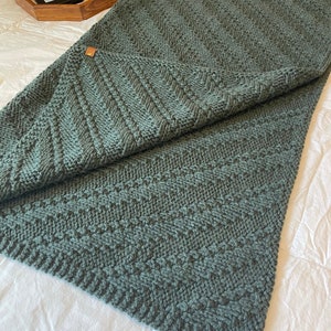 May include: A soft, green knitted throw blanket with a textured pattern. The blanket is folded in half and lying on a white surface.