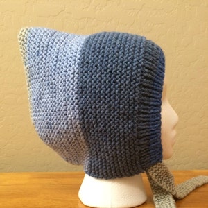 May include: A blue and gray knitted baby bonnet with a long matching tie. The bonnet is on a white mannequin head.