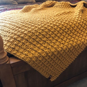 May include: A mustard yellow knitted throw blanket with a textured pattern. The blanket is draped over a wooden bed frame.