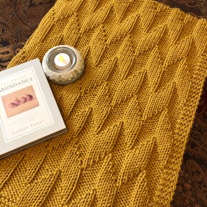 May include: A mustard yellow knitted throw blanket with a cable knit pattern. The blanket is folded and lying on a patterned surface.