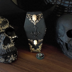 May include: Black coffin-shaped ring holder with gold accents and a cross design. The holder is decorated with small skull and bone details. A green ring is displayed on the holder.