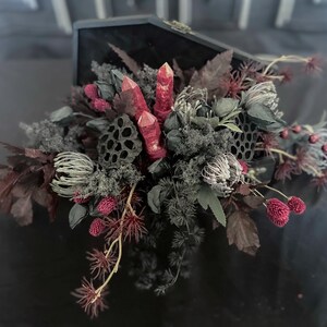 May include: A dark, open coffin filled with a floral arrangement. The arrangement features red crystals, dried flowers, and seed pods in shades of gray, burgundy, and green. A unique gothic home decor item.