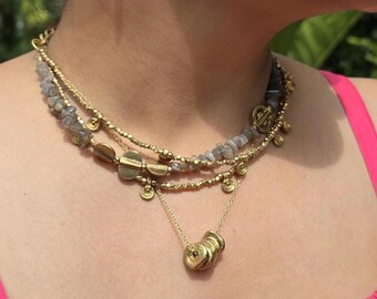 May include: A multi-strand necklace set with three layers. The top layer has grey stone beads, the middle has gold-tone metal discs, and the bottom has a pendant with three stacked gold-tone rings. The necklace is worn over a pink top.