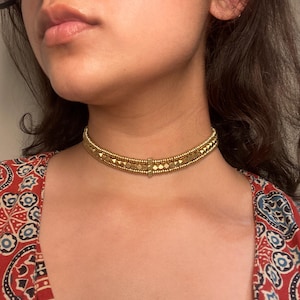 Puede incluir: Un collar gargantilla dorado con un diseño geométrico. El collar presenta pequeñas cuentas redondas y detalles cuadrados y rectangulares. La gargantilla se lleva alrededor del cuello. La persona lleva una blusa estampada roja.