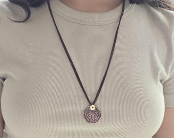 May include: A necklace featuring a dark brown cord and a circular pendant with a textured, hammered metallic surface. The pendant is attached to the cord by a small, smooth gold-toned circular connector, creating a simple and clean design.