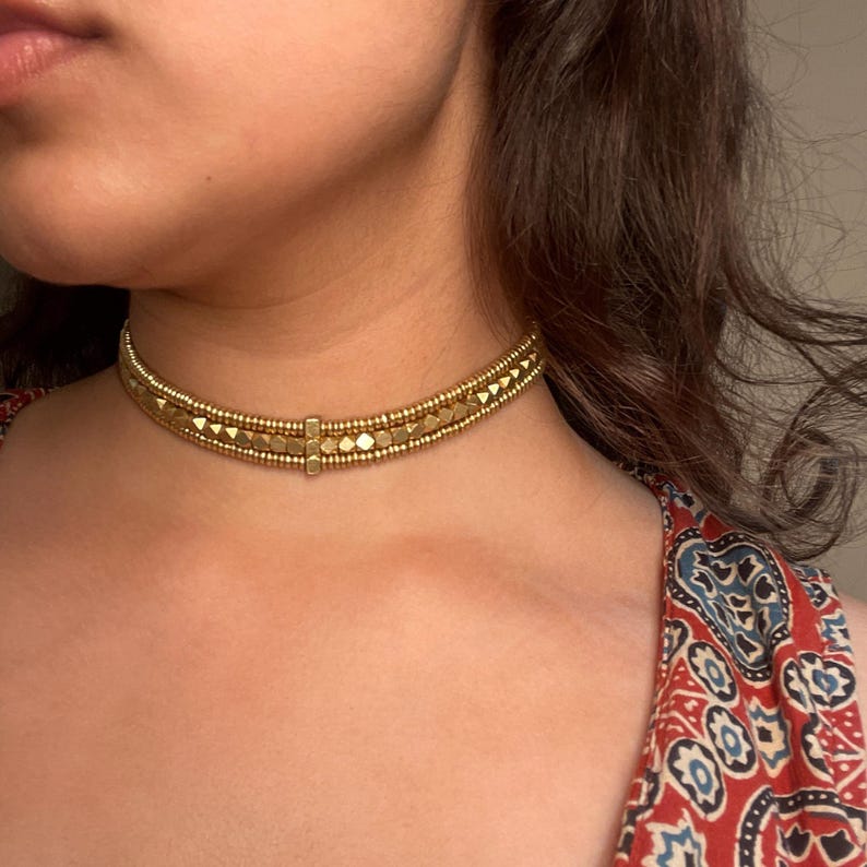 May include: Gold-toned choker necklace with a geometric design. The necklace features rows of small beads and larger, square-shaped elements. The choker is worn around the neck, and the background includes a patterned red and blue fabric.