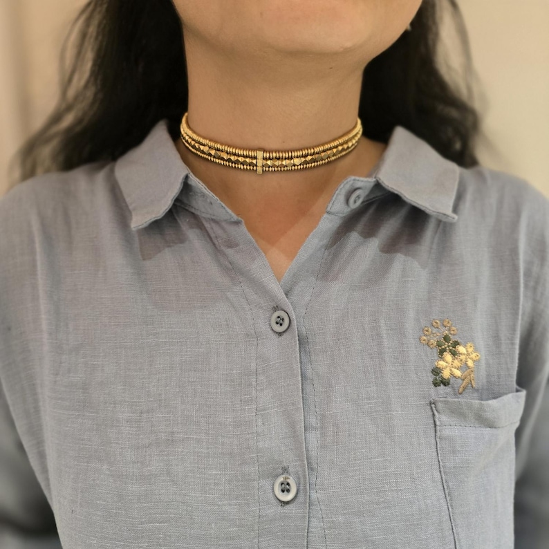 May include: A gold-colored choker necklace with three rows of beaded design. The necklace is worn around the neck and is the focal point of the image. The person is wearing a light blue button-down shirt.