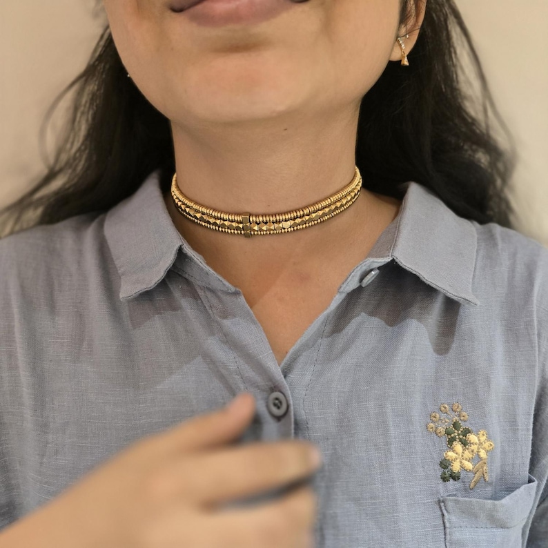 May include: A gold choker necklace with three strands of beads and geometric shapes. The necklace is worn on a person wearing a light blue button-down shirt. The shirt has a floral embroidery on the pocket.