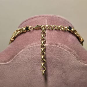 May include: Close-up of a gold-toned necklace with a beaded design and black accents. The necklace features a chain extender for adjustable sizing. The clasp and chain are visible, showcasing the intricate details of the jewelry.
