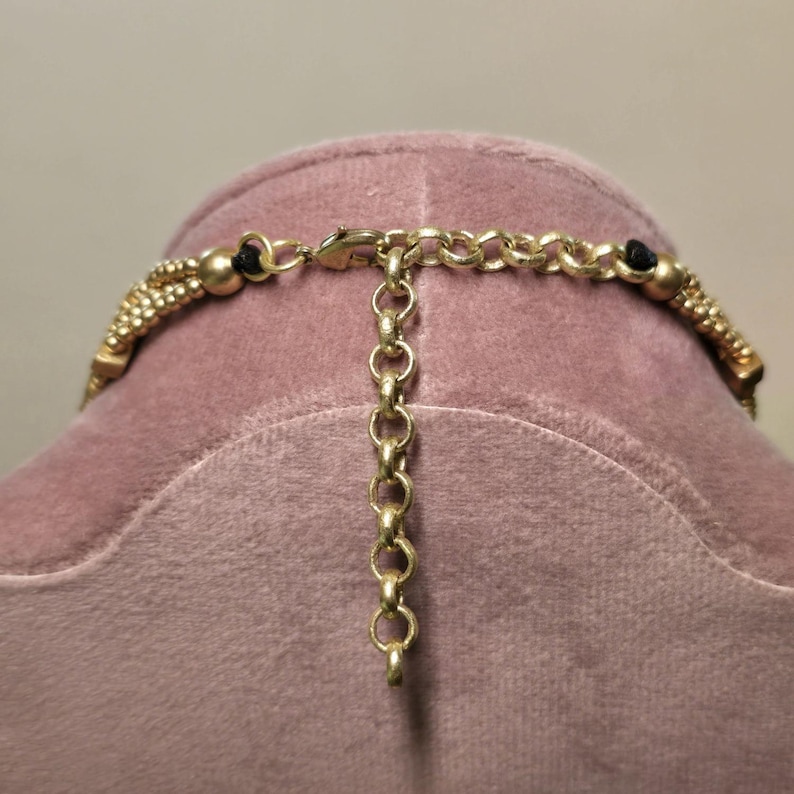 May include: Close-up of a gold-toned necklace with a beaded design and black accents. The necklace features a chain extender for adjustable sizing. The clasp and chain are visible, showcasing the intricate details of the jewelry.